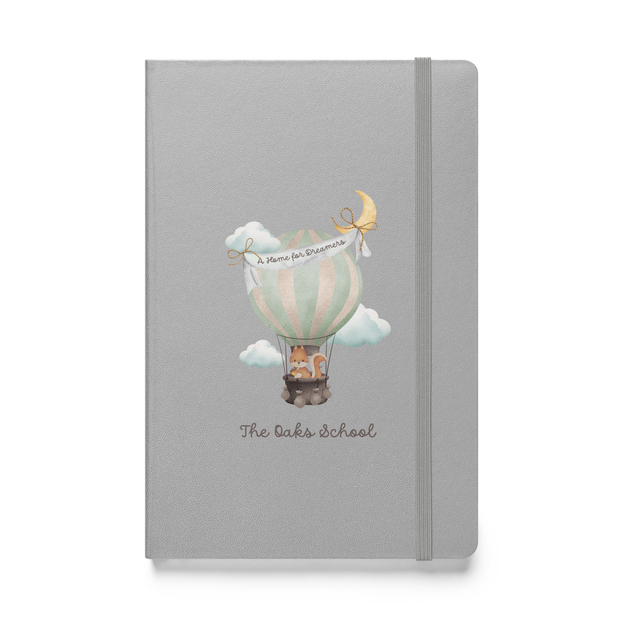 Product mockup: A silver colored journal on a white background featuring a hot air balloon containing a squirrel and text that says "The Oaks School" and "A Home for Dreamers."